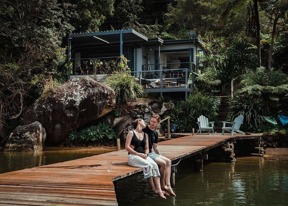 A couple sitting on a private jetty with a light blue cottage in the background